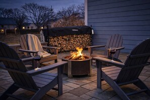 A fire pit surrounded by Adirondack chairs, a covered grill, and a stone wall with logs.