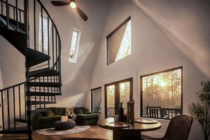A modern interior with a spiral staircase, a green sofa, a round table, and large windows with a forest view. - Ambrosia Abode | Grand Opening (Hendersonville)