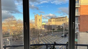 Vista desde una ventana a una calle de la ciudad, con un edificio histórico, un edificio moderno y un árbol en primer plano.