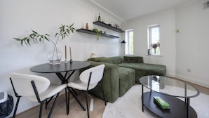 Una sala de estar moderna con una mesa redonda de vidrio, dos sillas blancas, un sofá verde y una mesa de centro de vidrio. Hay dos estantes flotantes sobre el sofá con objetos decorativos.