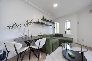 A modern living room with a round glass table, two white chairs, a green sofa, and a glass coffee table. There are two floating shelves above the sofa with decorative items. - Central 2BR Flat w/balcony Near Temple Bar (The Liberties)