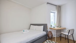 Una cama bien tendida con sábanas blancas, un escritorio con una silla y una ventana con cortinas.