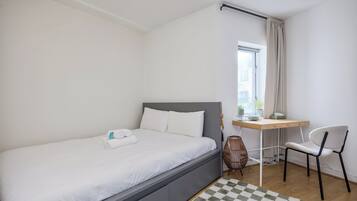 A neatly made bed with white linens, a desk with a chair, and a window with curtains.