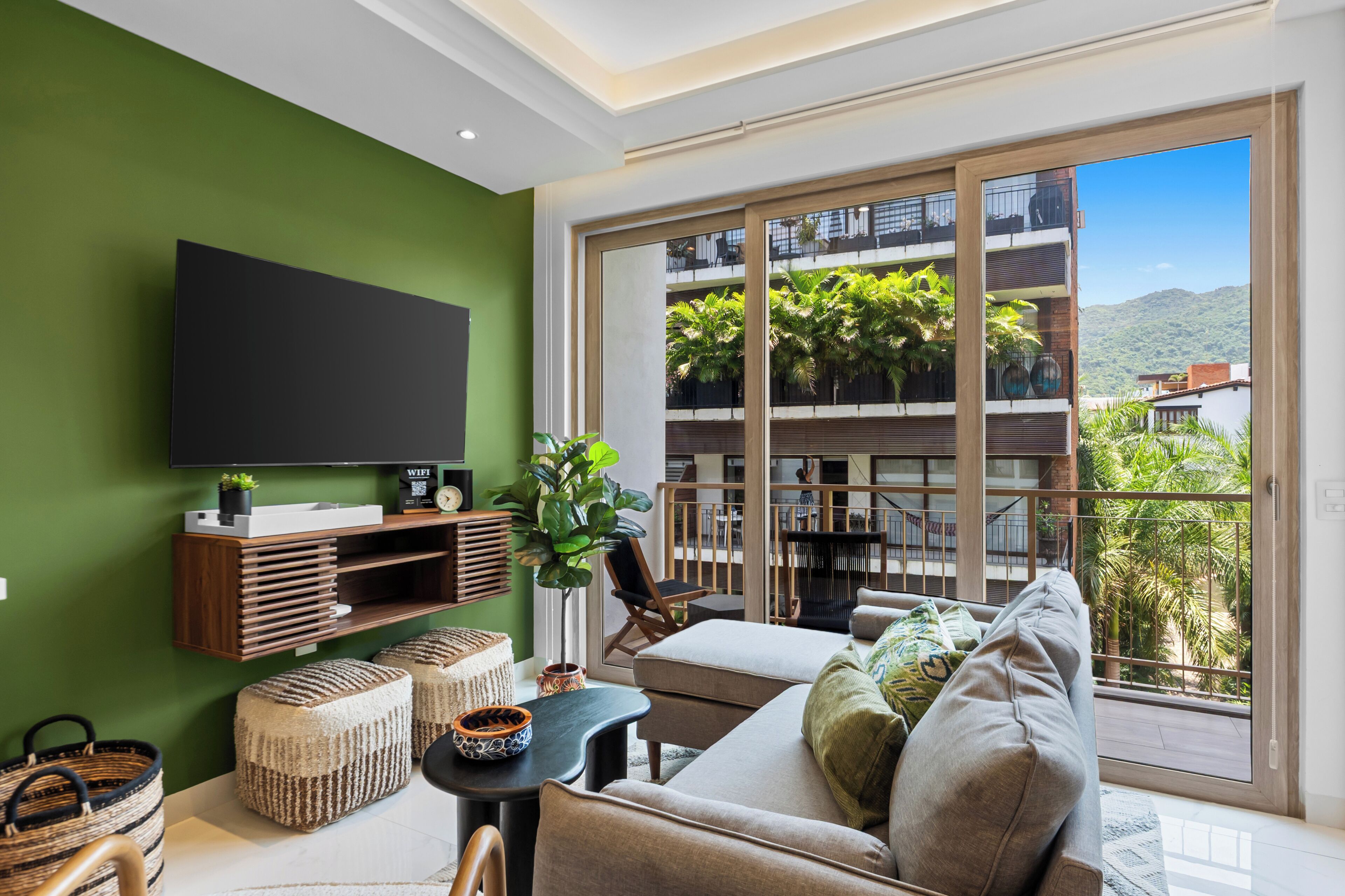 Un salon moderne doté d’un téléviseur à écran plat, d’un canapé, d’une table basse et d’un balcon offrant une vue sur la verdure et les bâtiments.