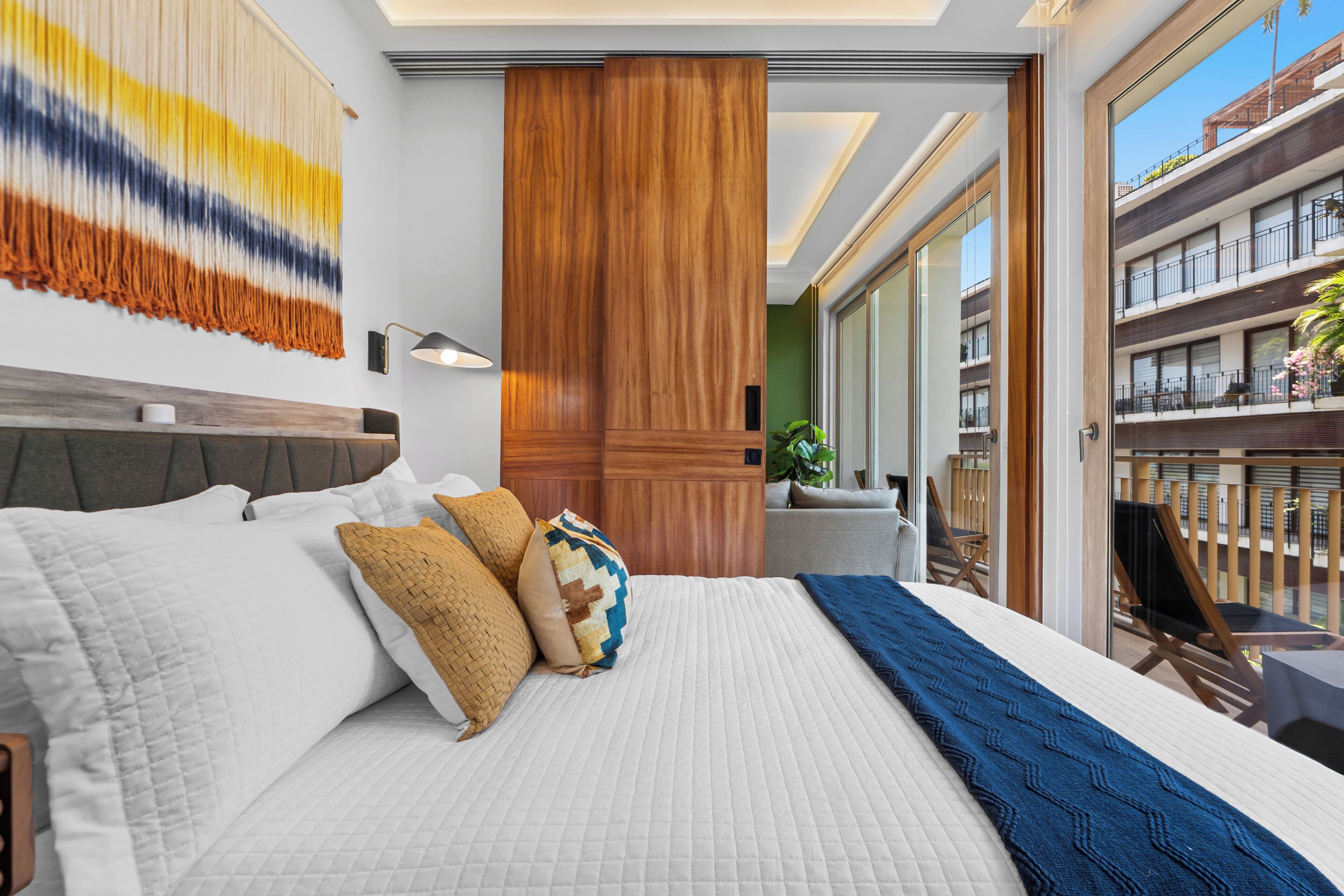 Habitación de hotel con una cama grande, cabecera de madera, lámpara de pared y vistas al balcón exterior.