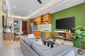 A modern living room with a grey sofa, a dining area with a table and chairs, a kitchen with wooden cabinets and a built-in oven, and a green accent wall with a mounted TV. - Luxury 2B Romantic Zone Condo , Rooftop Pool, 2-Min Walk to Beach (Puerto Vallarta)