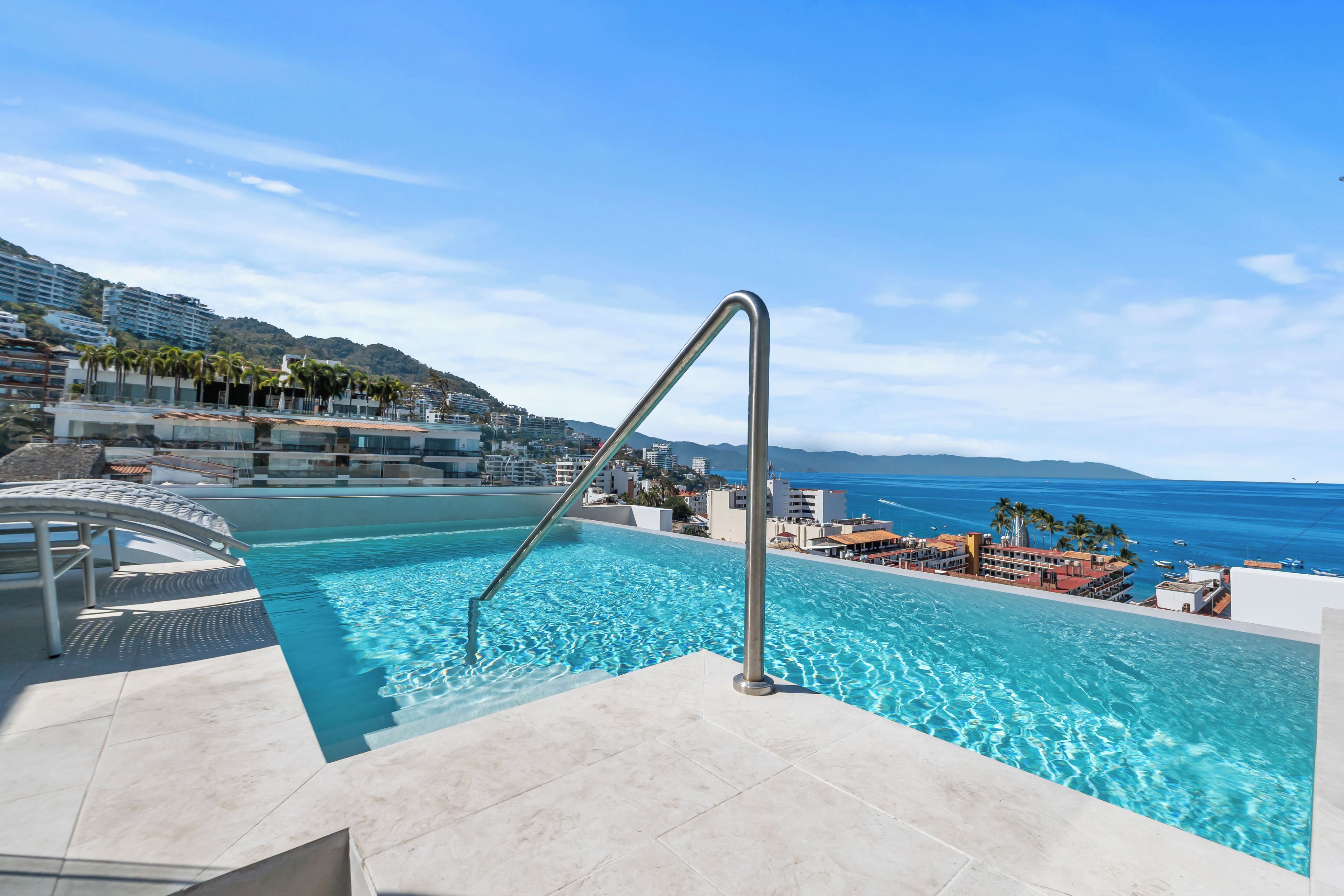 Une piscine avec un escalier en métal, entourée d’une terrasse carrelée, donnant sur une zone côtière avec des bâtiments et un ciel dégagé.