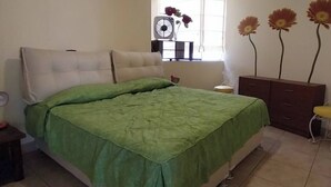 Ein Schlafzimmer mit einem Bett, einem Nachttisch, einer Kommode und einem Fenster mit Ventilator.