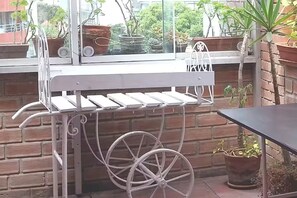 Un área de estar exterior de metal con una mesa y sillas, rodeada de plantas en macetas y un muro de ladrillo.