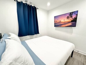 Ein Schlafzimmer mit Bett, Vorhängen, einem an der Wand befestigten Fernseher und einem Sonnenuntergangsbild.