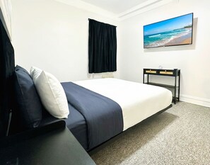 Ein Hotelzimmer mit Bett, Nachttisch in Schwarz, Flachbildfernseher, schwarzem Vorhang und einem Gemälde mit Strandblick.