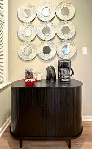 A black cabinet with a coffee maker and a kettle on top, and a red bowl with snacks beside it. - Cozy and Charming Forest Park Apt with Parking - Easy Access to City + Airports (Forest Park)