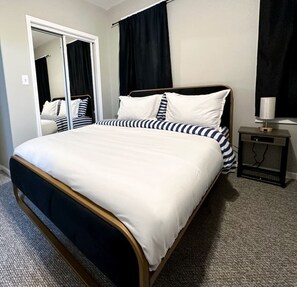 A bed with a white and navy striped bedspread, a wooden headboard, and a bedside table with a lamp. - Cozy and Charming Forest Park Apt with Parking - Easy Access to City + Airports (Forest Park)