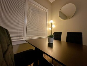 A dining area with a table, chairs, a potted plant, a wall-mounted mirror, and a window with blinds. - Cozy and Charming Forest Park Apt with Parking - Easy Access to City + Airports (Forest Park)