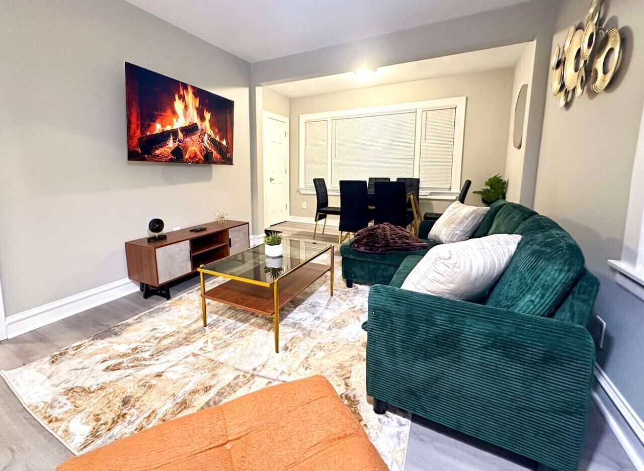 A living room with a green sofa, a glass coffee table, a fireplace with a fire, and a dining area in the background.