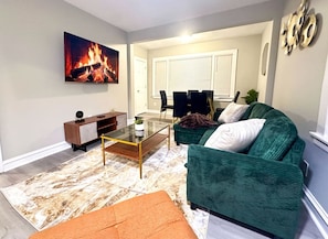 Ein Wohnzimmer mit einem grünen Sofa, einem Glastisch, einem Kamin mit Feuer und einem Essbereich im Hintergrund.