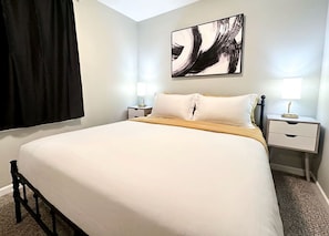 Ein Hotelzimmer mit einem Bett, zwei Nachttischen, einem an der Wand hängenden abstrakten Gemälde und einem Fenster mit Vorhängen.