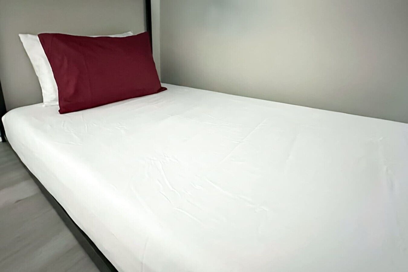A single bed with a white mattress and a red pillow in a room with a plain wall and a light-colored floor.