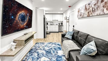 A modern living room with a large flat-screen TV, a black leather sofa, a blue patterned rug, and a galaxy-themed artwork on the wall.
