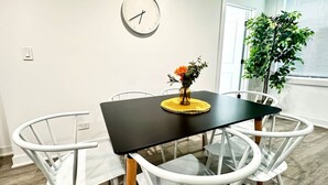 Une table à manger avec un plateau noir et des chaises blanches, une horloge au mur et une plante près de la fenêtre.