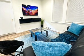 Ein Wohnzimmer mit einer Couch, einem Couchtisch, einem an der Wand befestigten Fernseher und einem Fenster mit Jalousien.