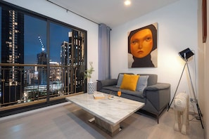 Ein modernes Wohnzimmer mit großem Fenster und Blick auf eine Stadtlandschaft, ein graues Sofa mit gelbem Kissen, ein Marmortisch und eine Stehlampe.