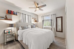 Ein Schlafzimmer mit einem Bett, einem Nachttisch, einem Deckenventilator, einem Fenster mit Vorhängen und einem Bücherregal.