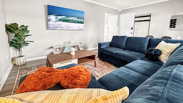 A living room with a dark blue sofa, a coffee table, a TV mounted on the wall, a potted plant, and a door with a coat rack.