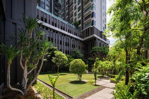A modern building with a courtyard featuring a well-maintained lawn, various trees, and a pathway. - 5 Min to Metro, Washer & Dryer, Essentials, Gym (Khet Khlong Toei)