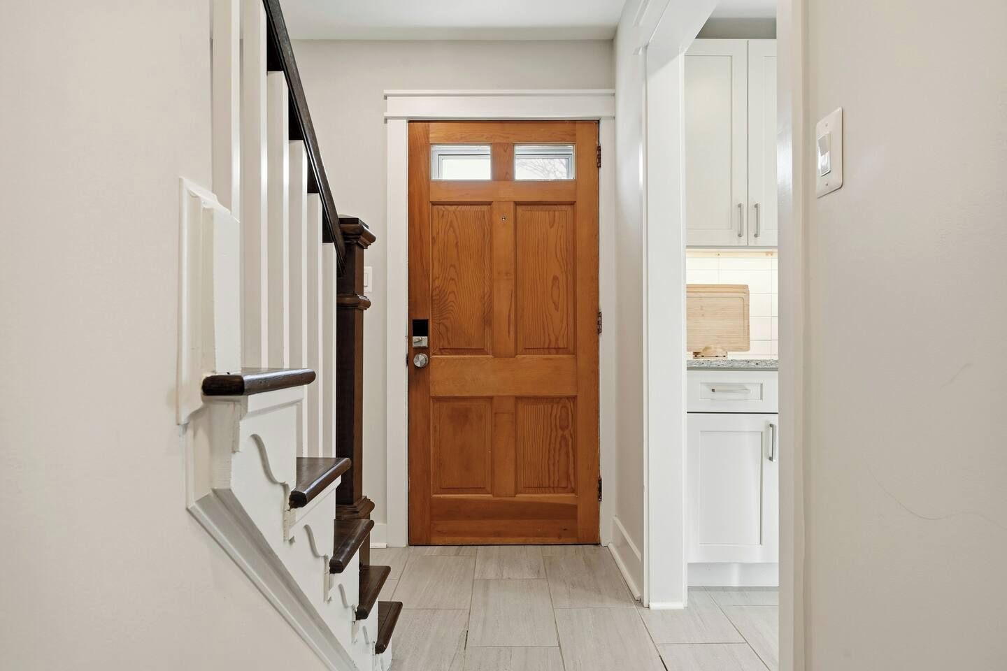 A staircase leading to a wooden door with a small window, white walls, and a kitchen area with white cabinets and a countertop.