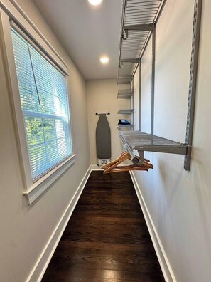 A narrow hallway with a wooden floor, metal shelving units, and a window with blinds. - Downtown Retreat with 2 Primary Suites (Leesburg)