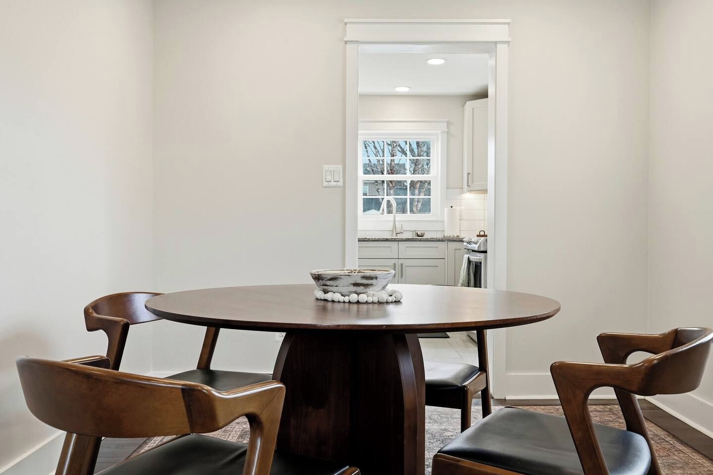 A dining area with a round table and four chairs, a bowl with decorative items on the table, and a view into a kitchen through an open doorway.