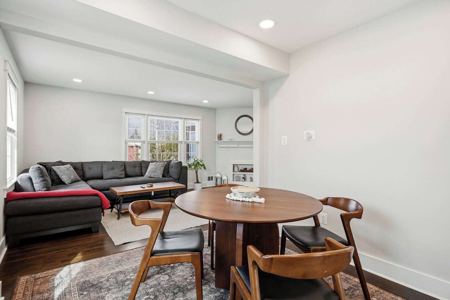 A modern living room with a dark grey sofa, a round wooden dining table with four chairs, and a large window providing natural light.