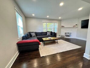 A living room with a dark wood floor, a black sectional sofa, a wooden coffee table, and a fireplace. - Downtown Retreat with 2 Primary Suites (Leesburg)