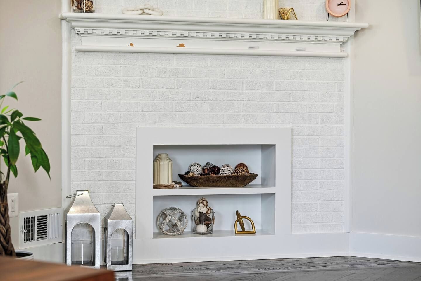 A white brick fireplace with decorative shelves above, a plant to the left, and a clock on the mantel.