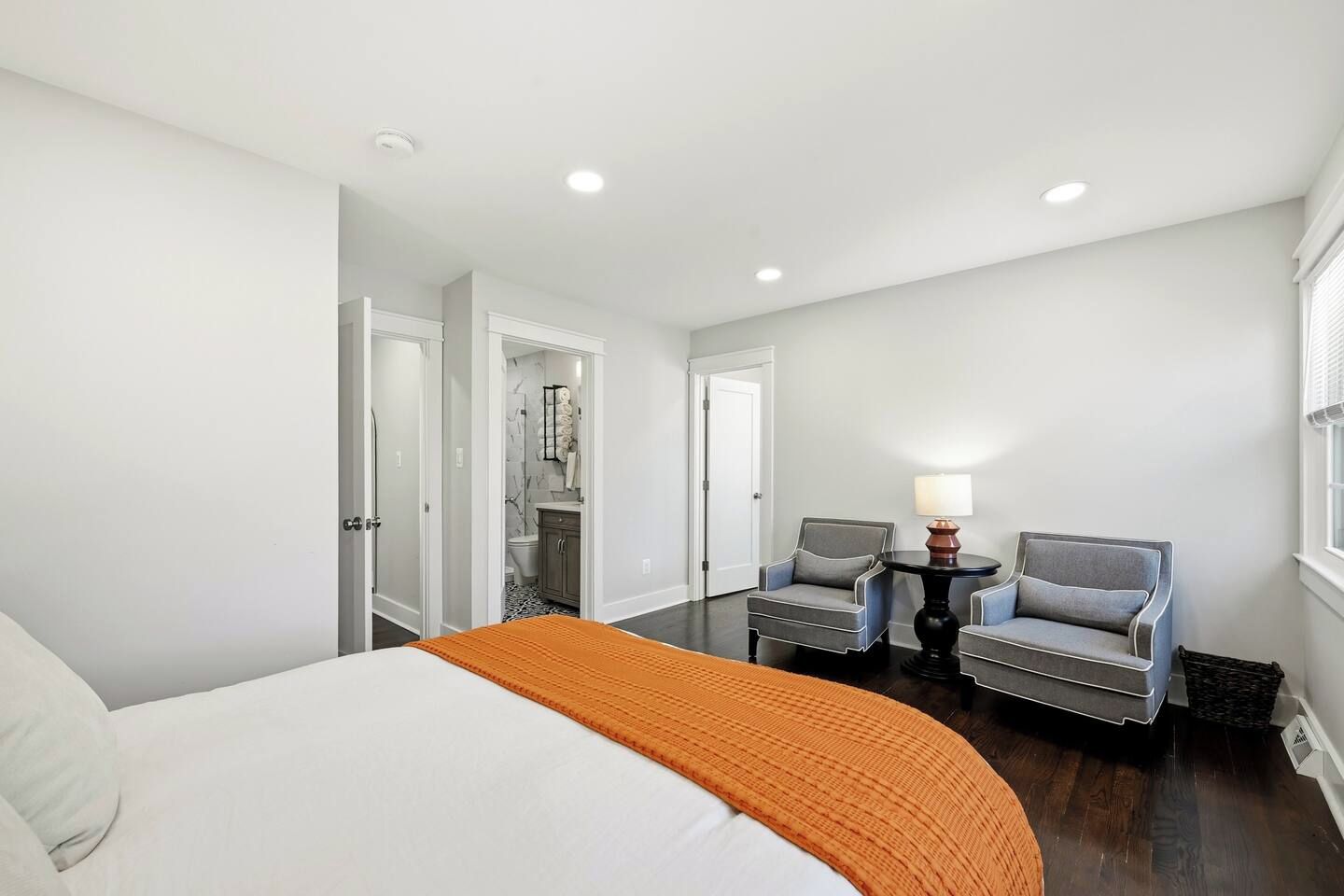 Une chambre d’hôtel avec un lit, deux fauteuils, une petite table avec une lampe et une salle de bain visible à travers une porte ouverte.
