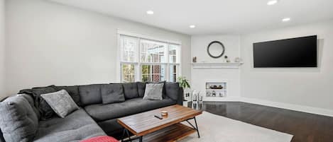 A modern living room with a sectional sofa, a coffee table, a fireplace, and a large flat-screen TV mounted on the wall.
