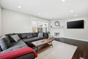 Een moderne woonkamer met een hoekbank, een salontafel, een open haard en een grote flatscreen televisie aan de muur.