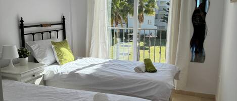 A hotel room with two beds, a nightstand, a lamp, a potted plant, and a view of the outside balcony.