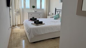 Een slaapkamer met een bed, nachtkastje, lamp, ventilator, een plant in pot en een raam met gordijnen.