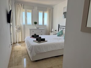 Une chambre à coucher avec un lit, une table de chevet, une lampe, un ventilateur, une plante en pot et une fenêtre avec des rideaux.