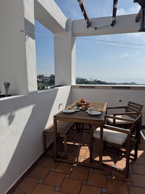 Un balcón con una mesa de madera preparada para el desayuno, vista a la ciudad y una pérgola con luces colgantes.