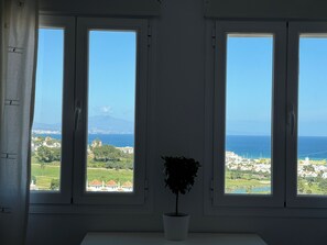 Une pièce dotée de trois fenêtres donnant sur un paysage côtier, une plante en pot sur le rebord de la fenêtre et un mur blanc.