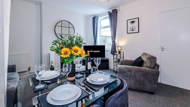 A dining area with a glass table set for two, a sofa, a TV, and a window with curtains. - Great 3-bed Home Perfect for Work Contractors (Ellesmere Port)