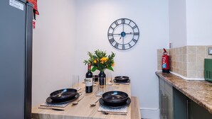A dining area with a wooden table set for four, a wall clock, and a fire extinguisher. - Stylish Home in Bolton Vibrant Central Location Sleeps 6 (Bolton)