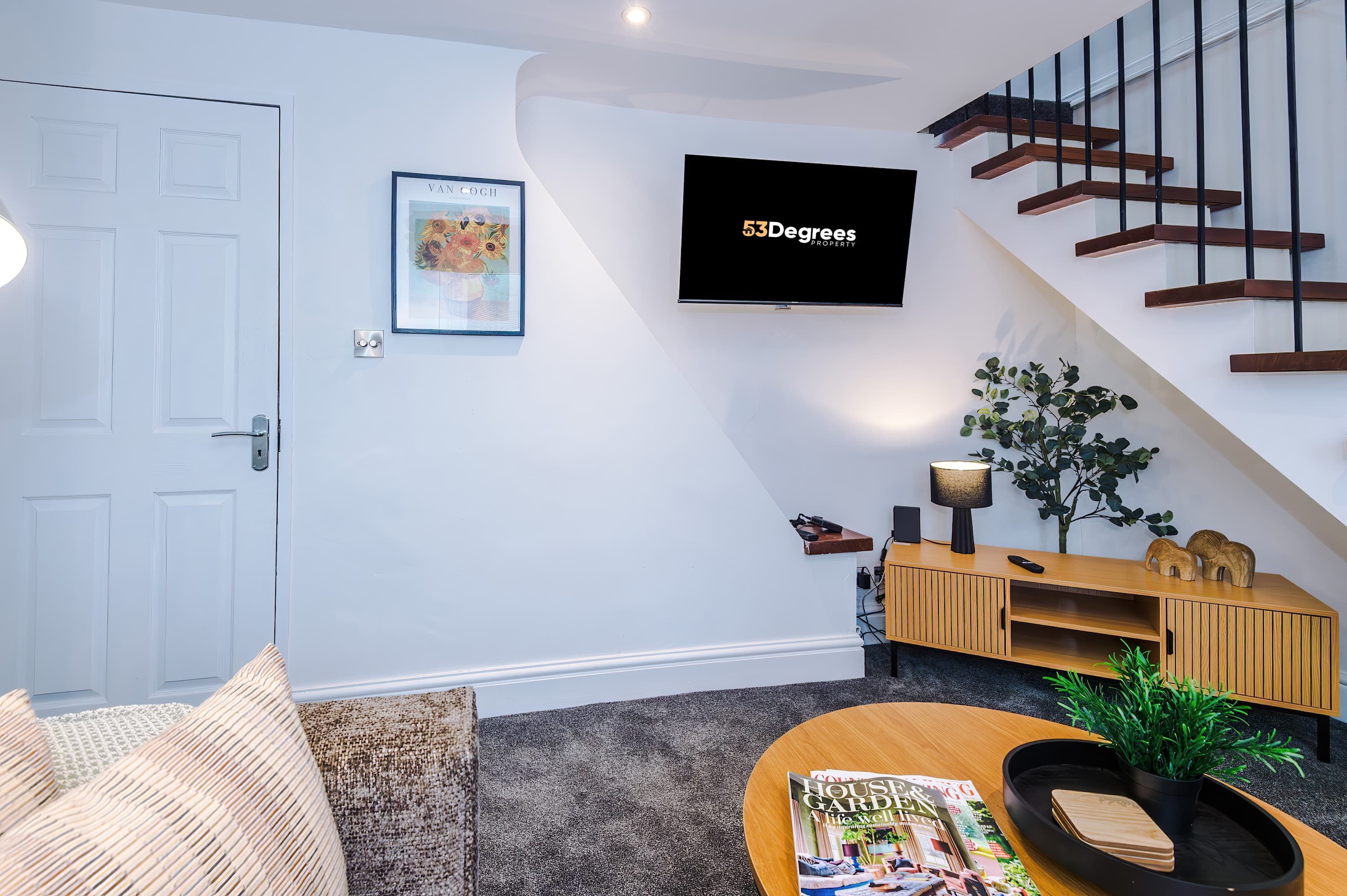 A modern living room with a flat-screen TV, a staircase, a sofa, a coffee table, and a bookshelf.