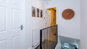 A hallway with white doors, a radiator, a black railing, and a woven wall hanging. - Stylish Home in Bolton Vibrant Central Location Sleeps 6 (Bolton)