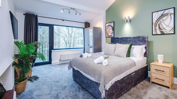 A bedroom with a large bed, bedside tables, a potted plant, and a view of the outdoors. - Stylish Home in Bolton Vibrant Central Location Sleeps 6 (Bolton)