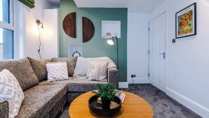 A living room with a sectional sofa, a coffee table with a plant, a wall-mounted lamp, and a framed artwork. - Stylish Home in Bolton Vibrant Central Location Sleeps 6 (Bolton)