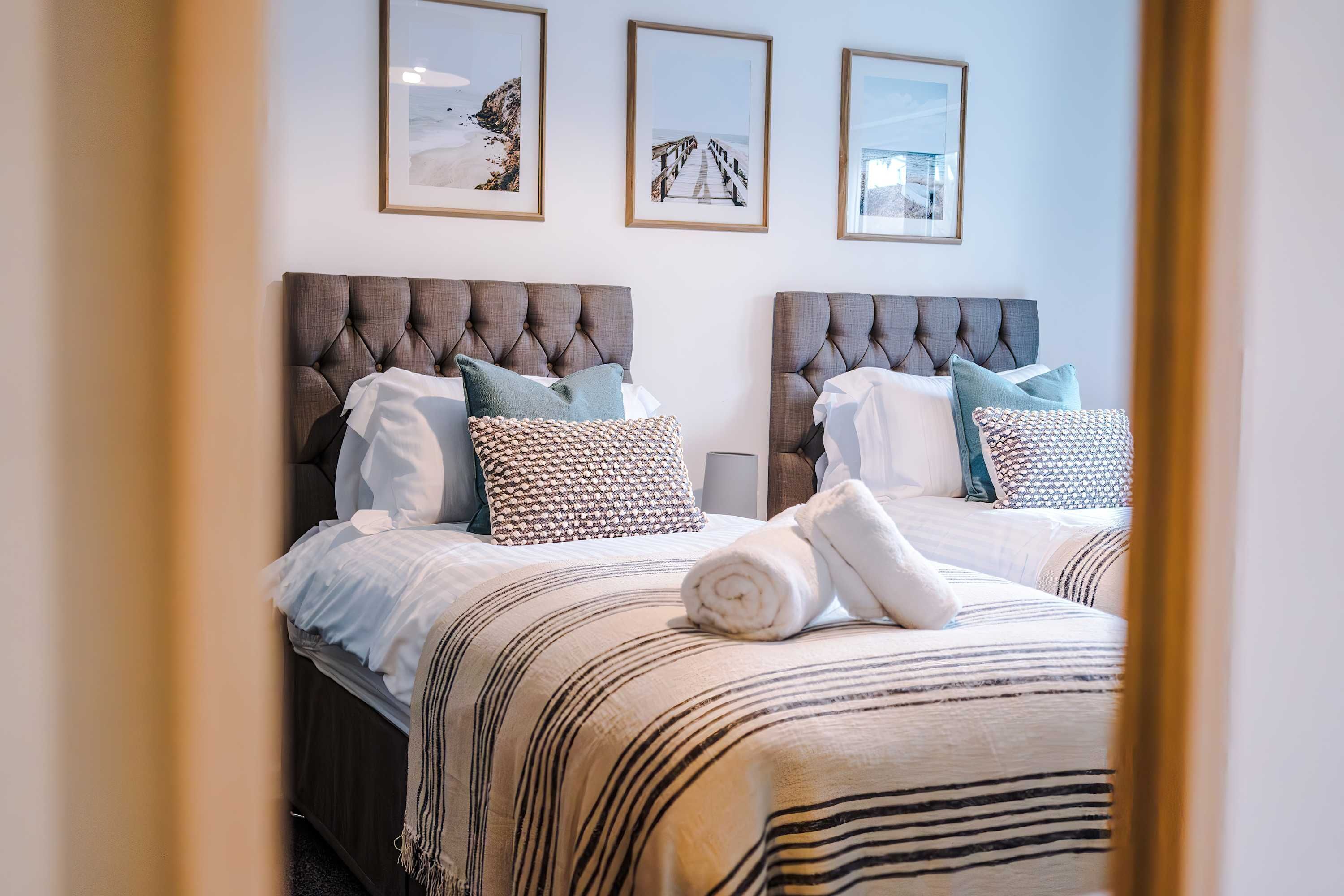 Une chambre d’hôtel avec deux lits, chacun doté d’une tête de lit capitonnée, de draps blancs et d’oreillers décoratifs. Des tableaux encadrés sont accrochés au mur au-dessus des lits.